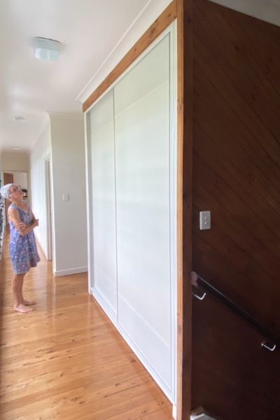 Homeowner admiring the transformed hallway linen cupboard fitted with modern white sliding doors, featuring white powder-coated mesh inserts at the top and bottom for airflow.