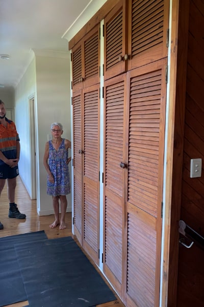 Homeowner inspecting old hinge louvre doors of a hallway linen cupboard that doesn't close properly, with a two-tiered design stretching floor to ceiling.