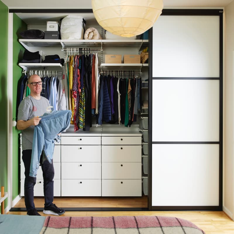 Spacious reach-in bedroom wardrobe with sliding doors revealing elegant Elfa Décor shelving, while a man thoughtfully organises his attire
