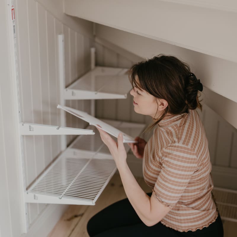 Resourceful use of space with a woman installing customisable Elfa shelving in a cozy under-stair nook to transform idle space into useful storage